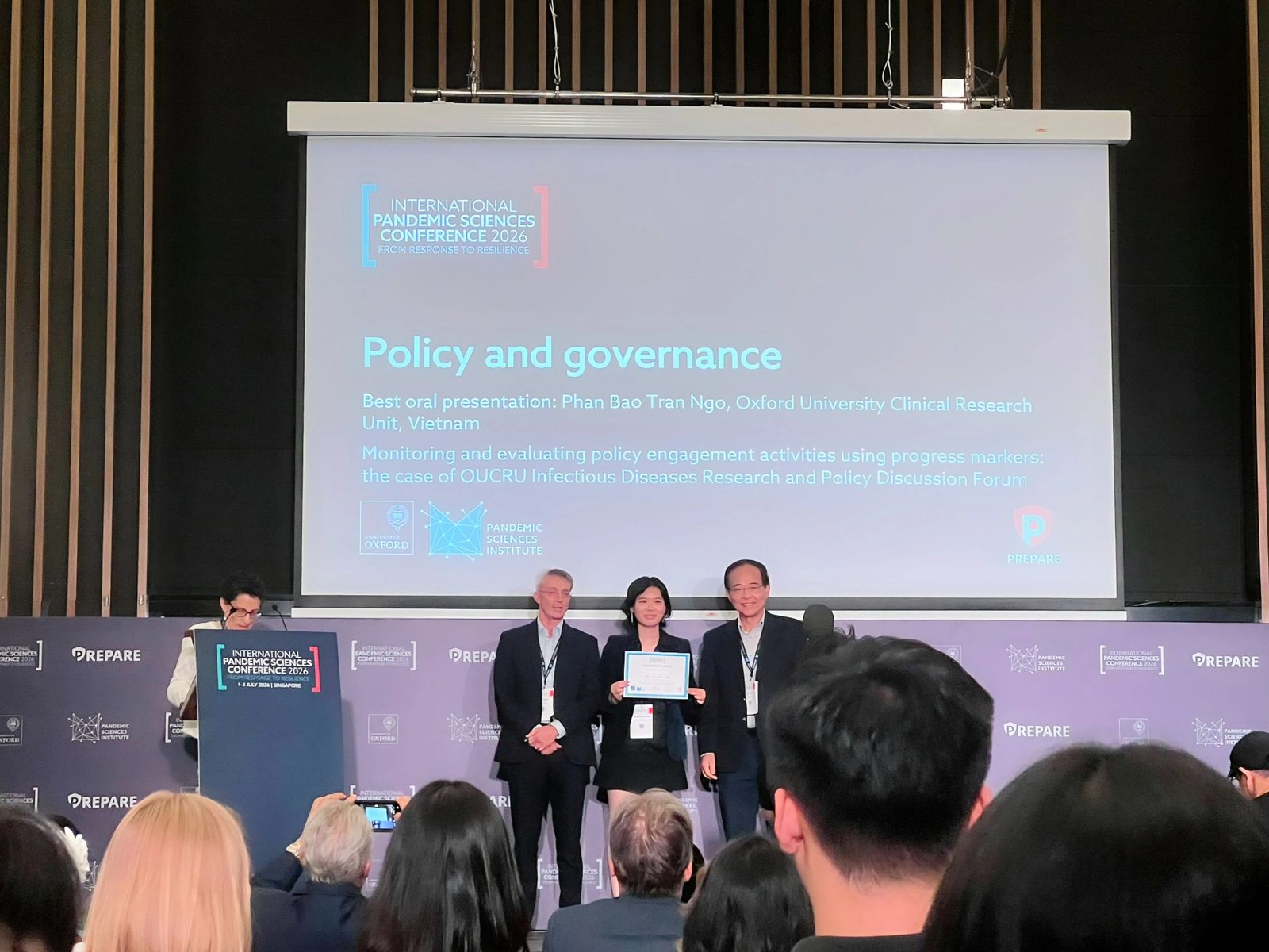 Ngô Phan Bảo Trân holds her award certificate on stage between two presenters at the International Pandemic Sciences Conference 2026 in Singapore, in front of a slide reading Policy and governance.